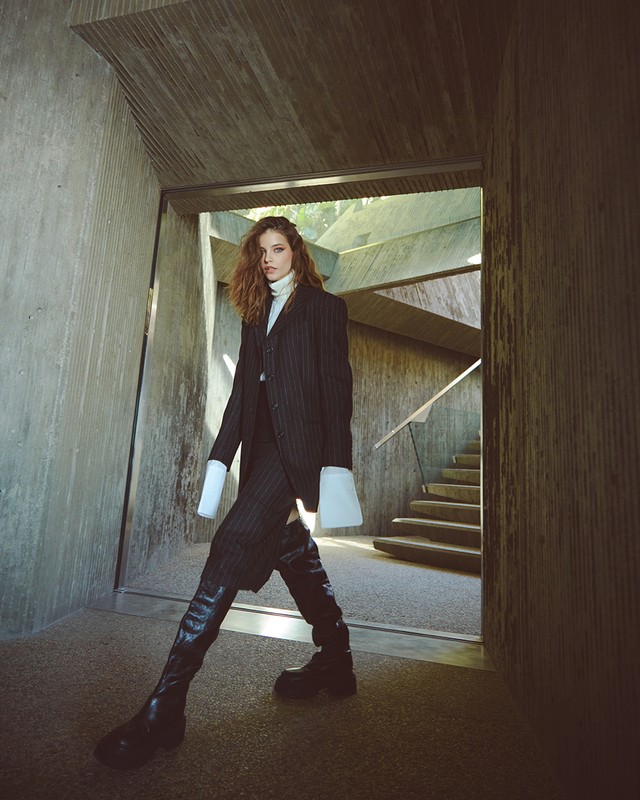 Fashion portrait of a woman in a pinstripe suit and black boots walking through a concrete hallway with stairs in the background.