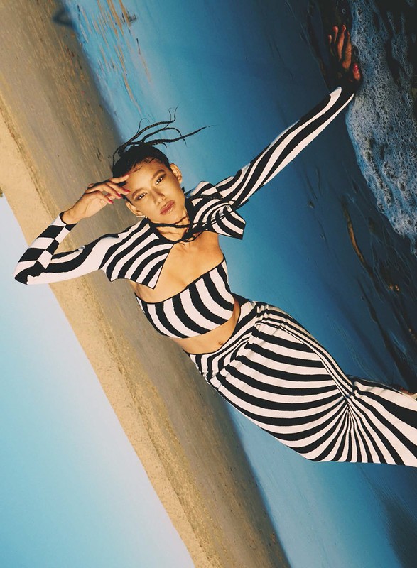 A woman poses in a black and white striped outfit against a beach and blue sky backdrop in a fashion or editorial shot.