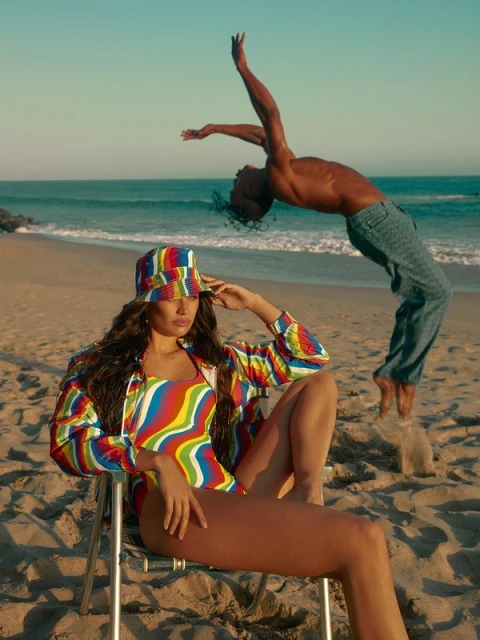 A woman in a colorful swimsuit sits on a beach chair while a man performs an acrobatic backbend on a sunny beach by the ocean.