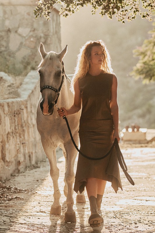 Woman in sleeveless dress walks with white horse on cobblestone road, lit by golden sunlight filtering through trees, nature.