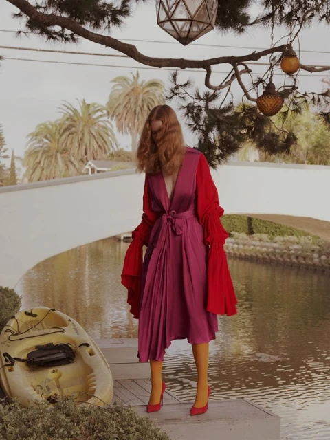 A woman in a red and magenta dress stands by a canal in a residential area, with palm trees and houses in the background.