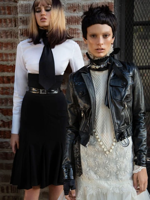 Two fashion models pose in front of a brick wall; one wears a white blouse with a black tie and skirt, the other a leather jacket and dress.
