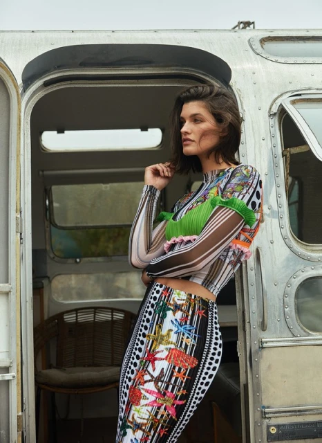 A woman with short dark hair poses in front of an Airstream trailer wearing a long-sleeved top and skirt with colorful patterns.