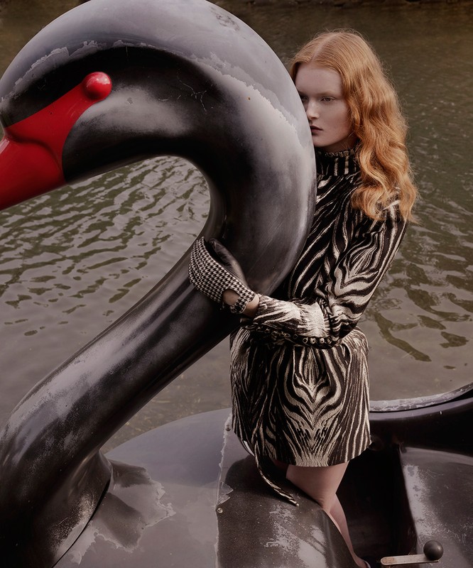 A red-haired woman in a zebra-print dress stands in a swan boat on a lake, fashion editorial photography with natural light.
