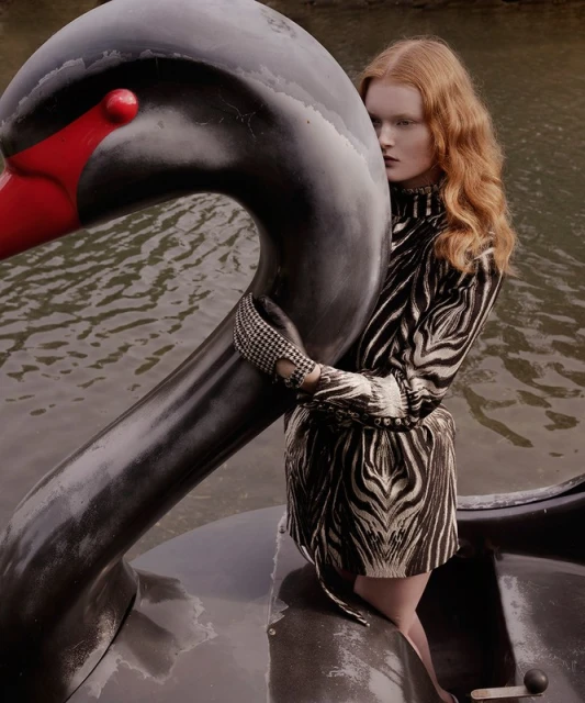 A red-haired woman in a zebra-print dress stands in a swan boat on a lake, fashion editorial photography with natural light.