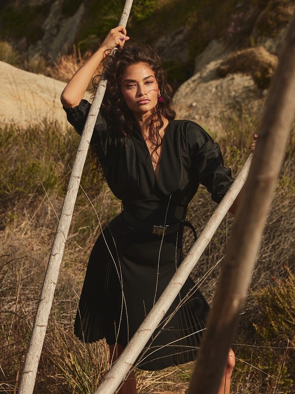 Brunette woman in black wrap dress poses with branches in an outdoor setting with grasses and rock formations, looking towards the camera.