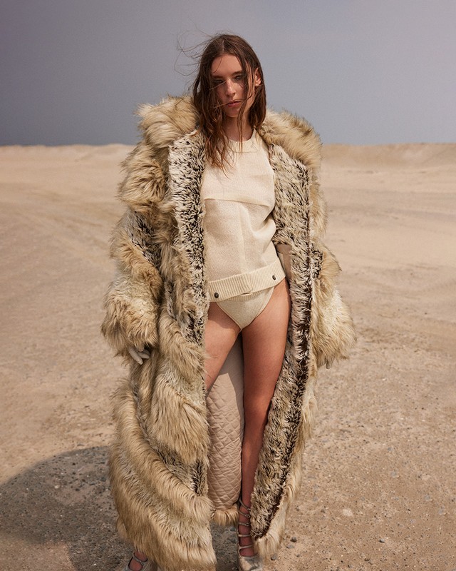 A woman with long brown hair poses in a sand dune wearing a tan knit outfit and a long fur coat against a light blue sky.
