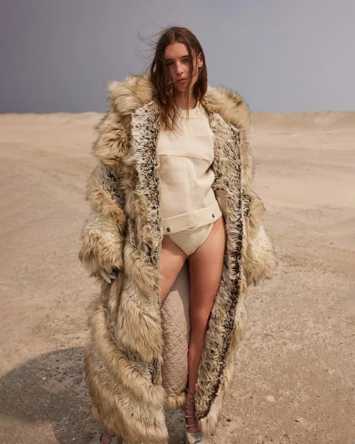 A woman with long brown hair poses in a sand dune wearing a tan knit outfit and a long fur coat against a light blue sky.