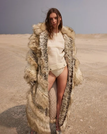 A woman with long brown hair poses in a sand dune wearing a tan knit outfit and a long fur coat against a light blue sky.