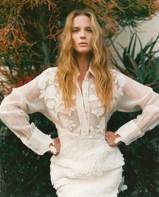 Fashion shot of a blonde adult woman in a sheer white floral-applique dress standing outdoors amid green and brown foliage, portrait view.