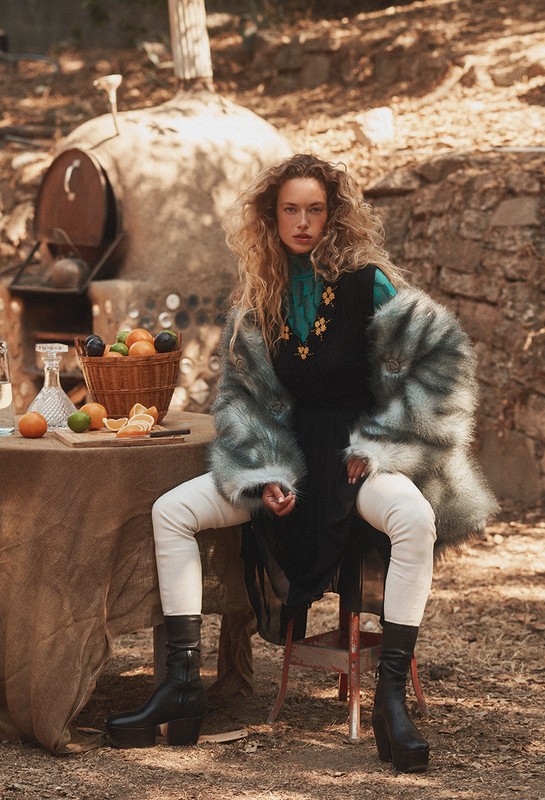 A fashionable woman sits on a chair outdoors wearing a faux fur coat, black dress, cream-colored leggings, and black platform boots in a wooded setting.
