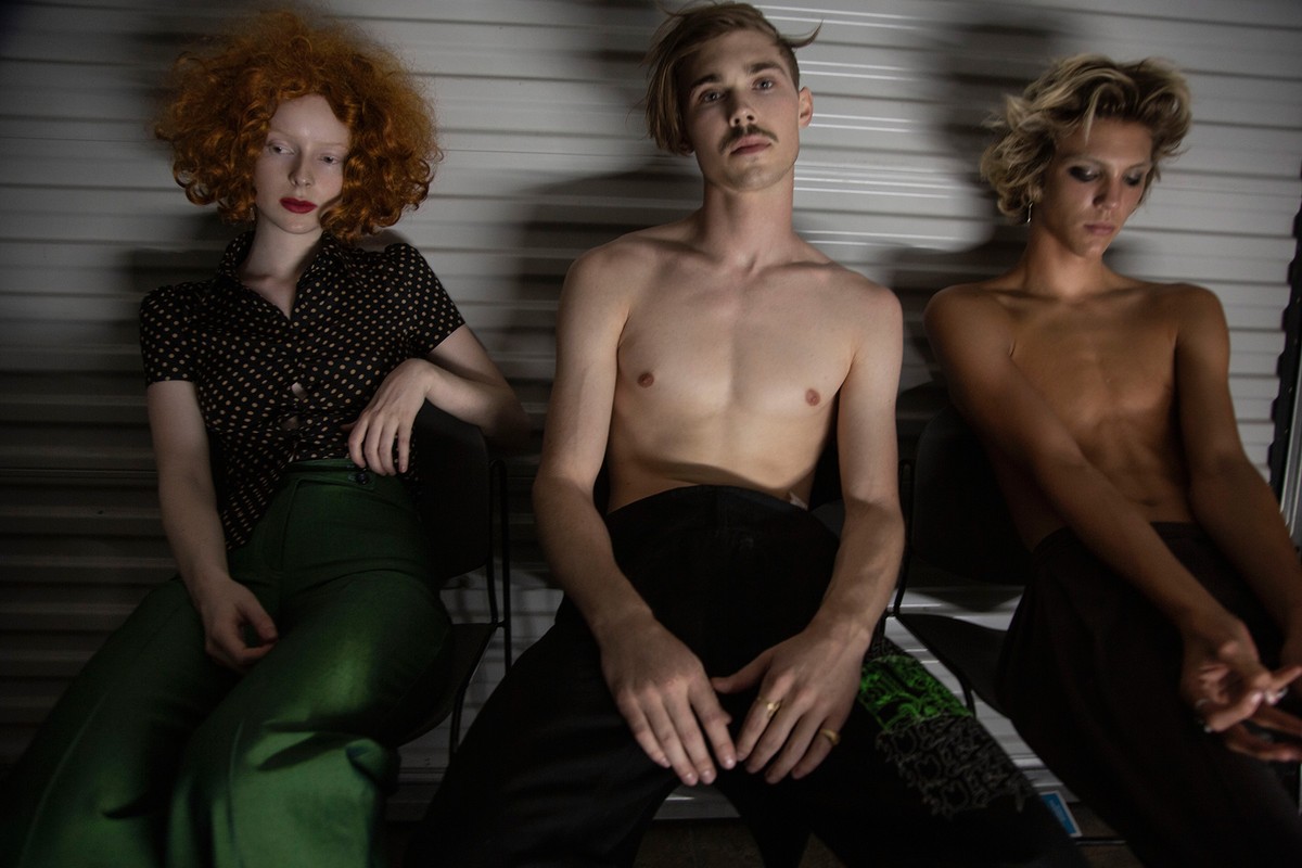 Three models seated indoors in a studio setting. One woman with red hair in a polka dot top, two men, one shirtless with fair skin.