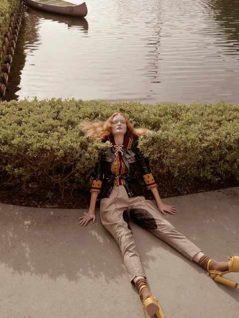 Redhead woman in fashionable jacket and trousers lies on a concrete path with greenery in the background near a river with a canoe.