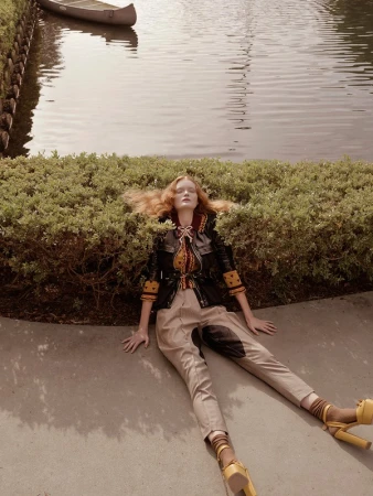 Redhead woman in fashionable jacket and trousers lies on a concrete path with greenery in the background near a river with a canoe.