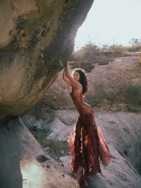 Woman in flowing dress poses artistically against a large rock in an outdoor landscape with sparse vegetation on a sunny day.