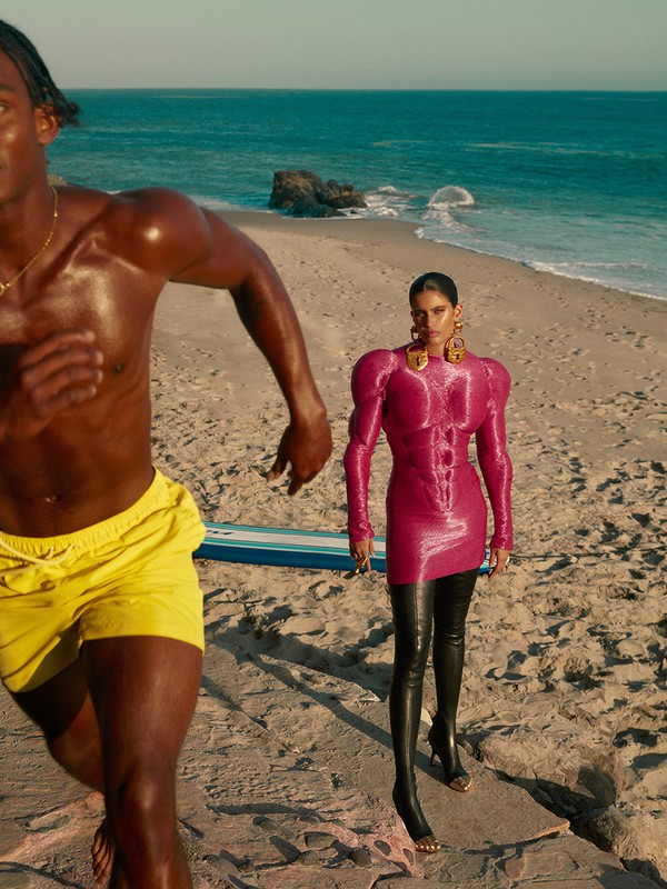 A man running on the beach next to a woman in a hot pink dress and black thigh high boots, blue sea and sky in the background.