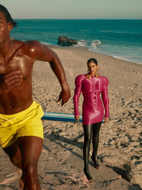 A man running on the beach next to a woman in a hot pink dress and black thigh high boots, blue sea and sky in the background.