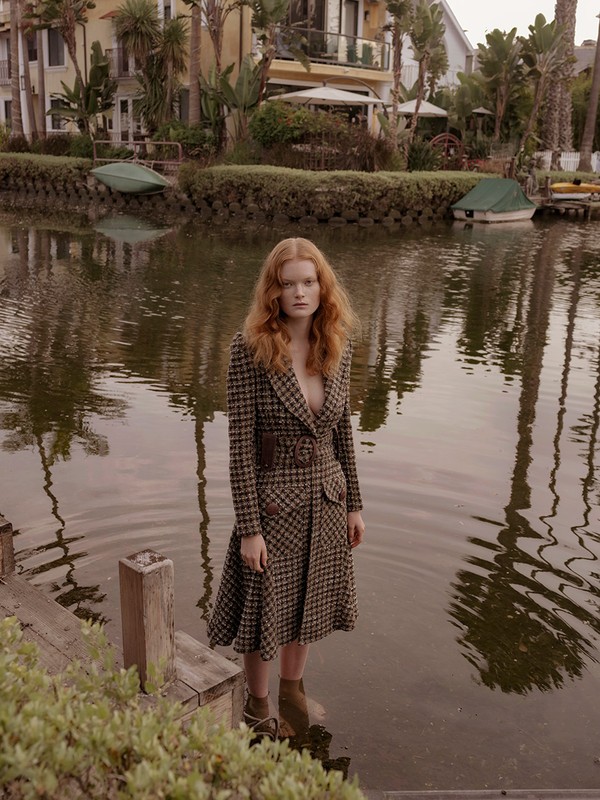 A woman with red hair and a patterned coat stands in the water of a canal lined with houses and trees in an outdoor setting.
