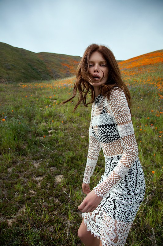 Adult woman wearing a white lace dress in a grassy field with orange flowers and green hills on a cloudy day in an outdoor fashion shot.
