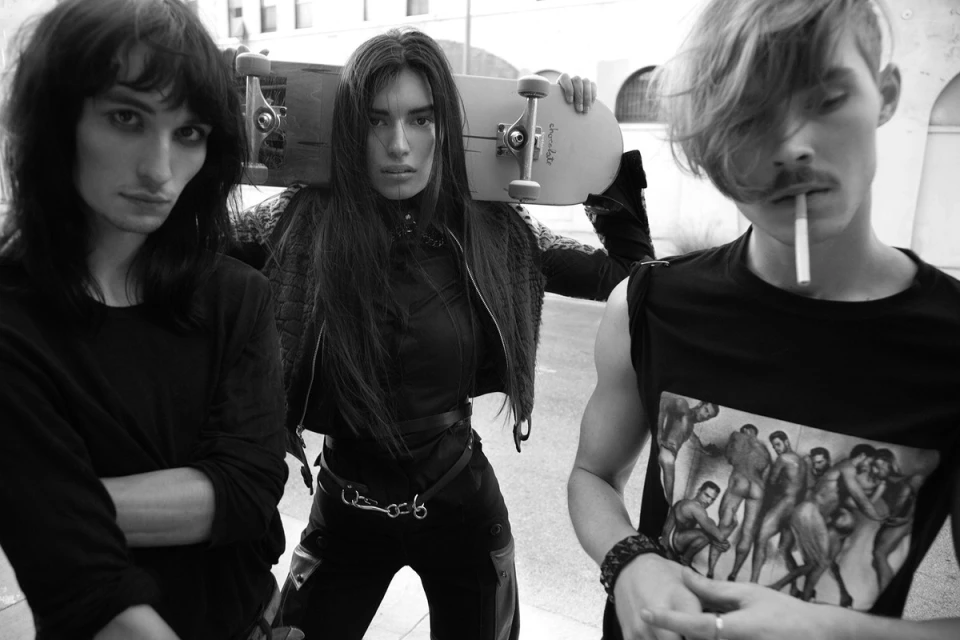 Black and white portrait of three edgy adults standing in an urban outdoor setting. A skateboard is held over one person's shoulders.