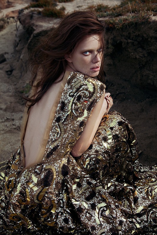 Young woman in a sequined dress sits on sand near rocks looking back at camera, facing forward with open back and arms crossed. 