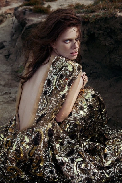 Young woman in a sequined dress sits on sand near rocks looking back at camera, facing forward with open back and arms crossed. 