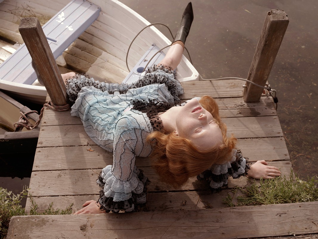 Red-haired woman lies on a wooden dock near a rowboat, wearing a ruffled dress and boots, eyes closed in a relaxed pose by the water.