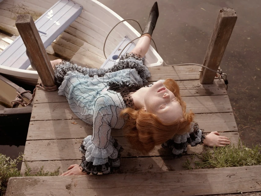 Red-haired woman lies on a wooden dock near a rowboat, wearing a ruffled dress and boots, eyes closed in a relaxed pose by the water.