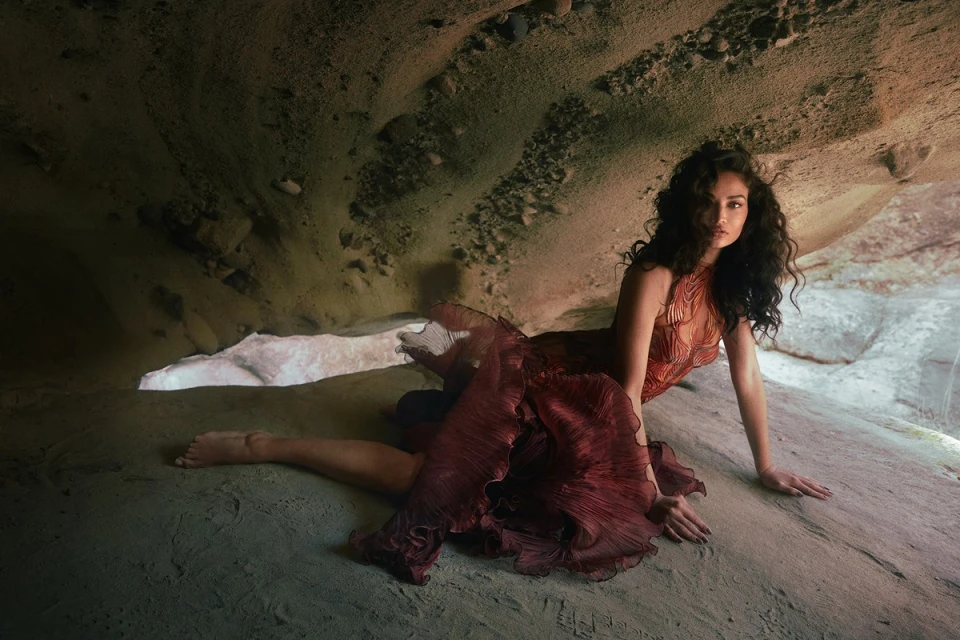 A woman in a maroon gown poses in a rock cave with sandy texture; the model is on her hands and knees looking at the camera.
