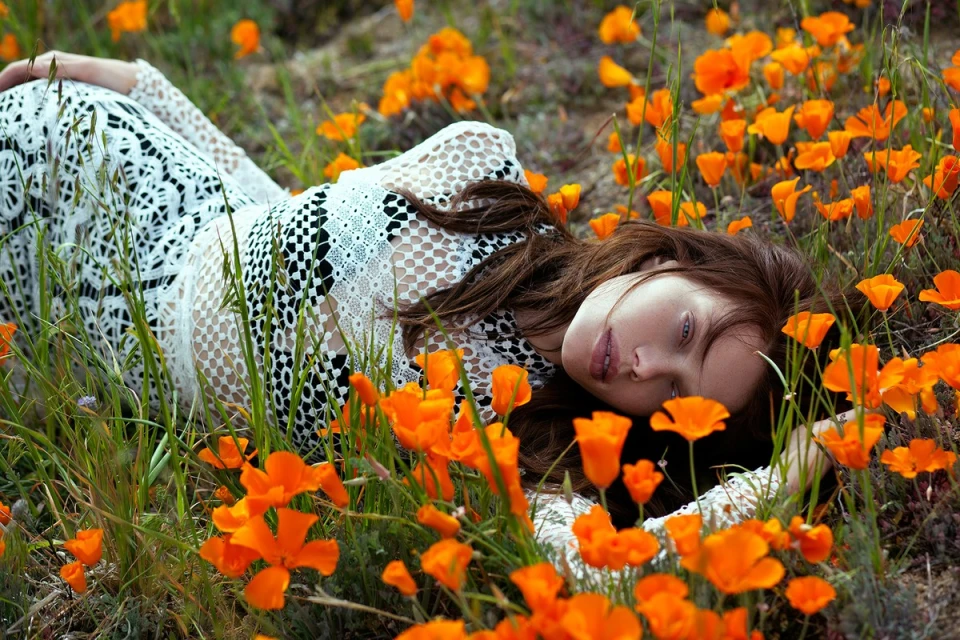 Woman in a white lace dress lies in a field of orange flowers, eyes closed, face turned toward the camera, shot outdoors.