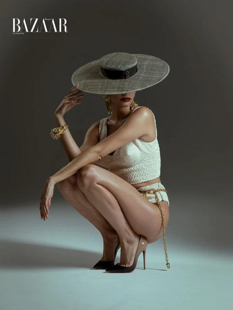 Fashion photography of a model in a light grey hat, tweed top and bottom, gold jewelry and black heels, posing in a squat position.