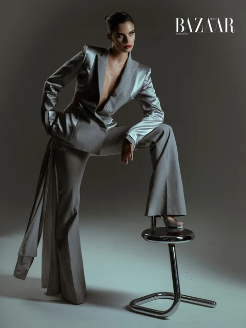 Fashion editorial shot of a model in a grey suit with leg posed on a chrome stool, studio lighting and a grey gradient backdrop.