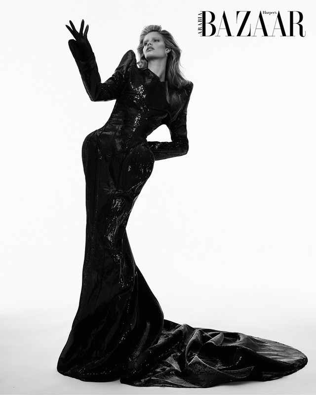 Editorial fashion shot in black and white. A woman in a long, shiny black dress and gloves poses in front of a white background.