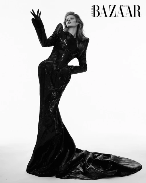 Editorial fashion shot in black and white. A woman in a long, shiny black dress and gloves poses in front of a white background.