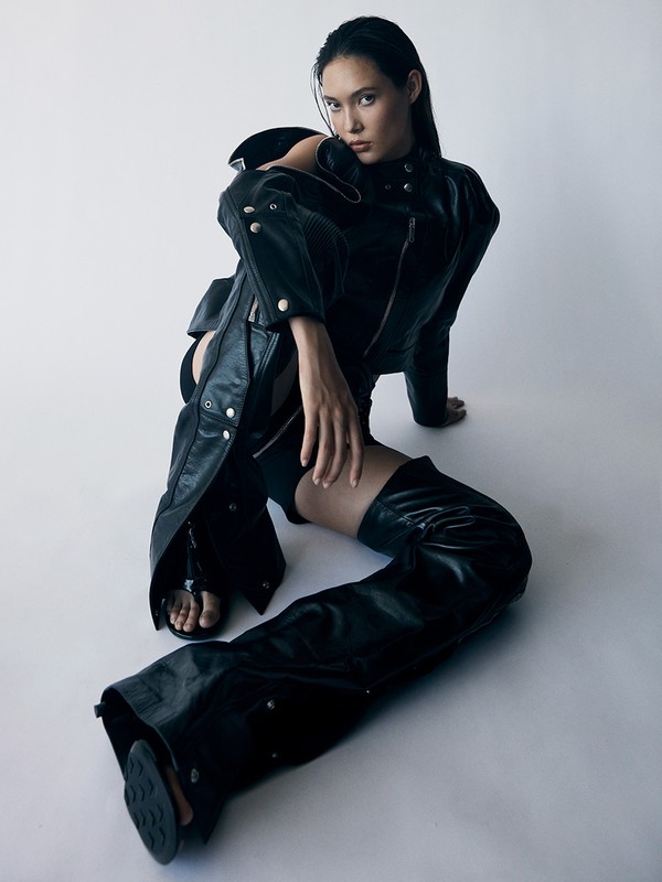 Fashionable young woman in a black leather jacket and chaps poses while sitting indoors against a white backdrop in a studio setting.