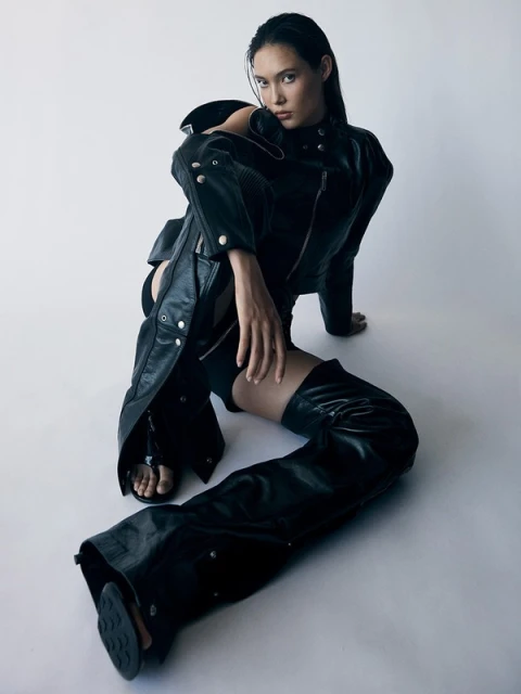Fashionable young woman in a black leather jacket and chaps poses while sitting indoors against a white backdrop in a studio setting.