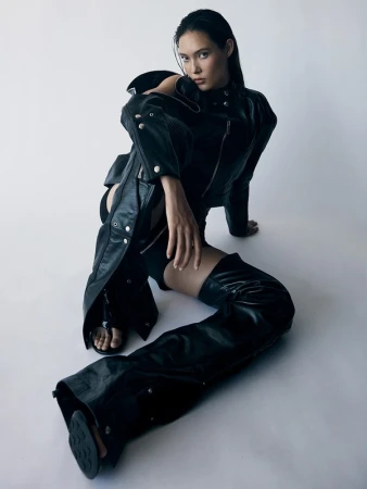 Fashionable young woman in a black leather jacket and chaps poses while sitting indoors against a white backdrop in a studio setting.