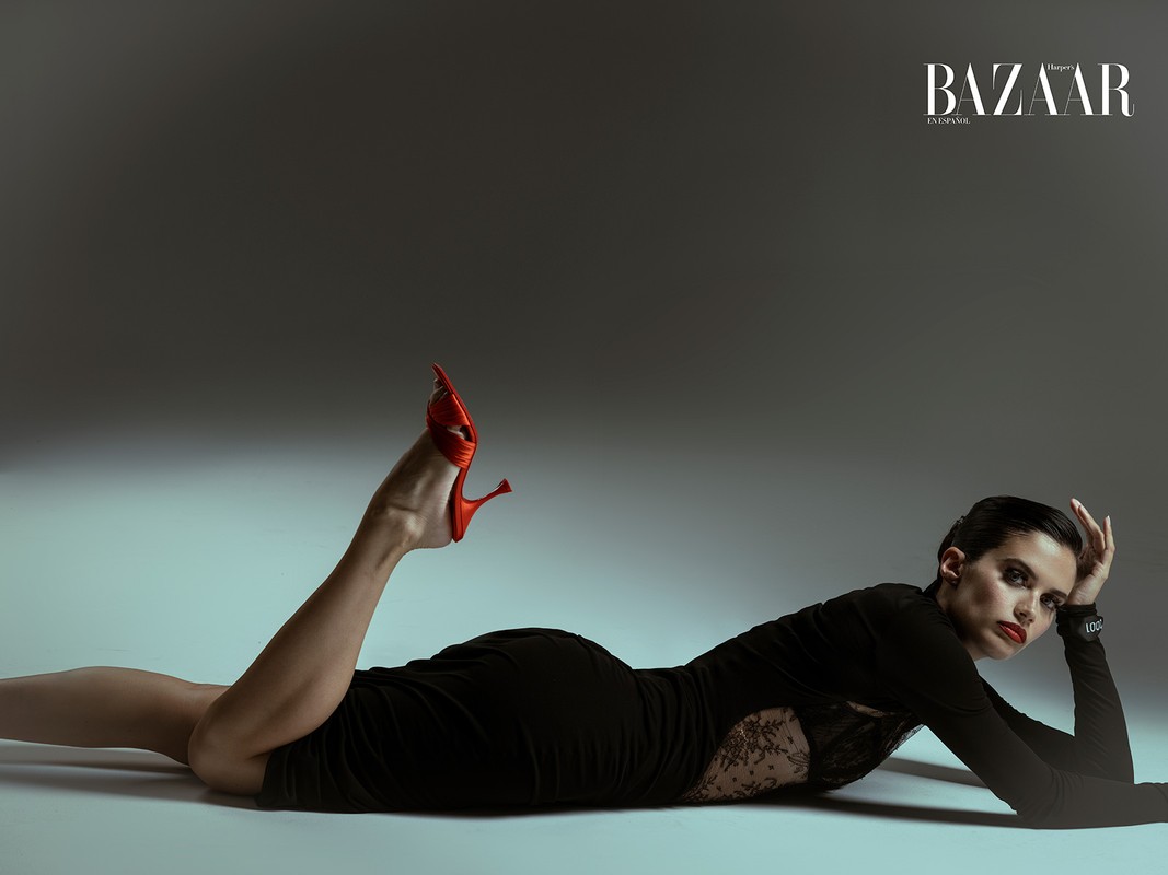 Fashion editorial shot of a woman in a black dress lying on a gray floor with red heels, Harper's Bazaar logo visible in the corner.
