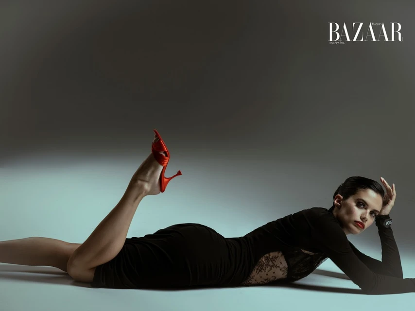 Fashion editorial shot of a woman in a black dress lying on a gray floor with red heels, Harper's Bazaar logo visible in the corner.
