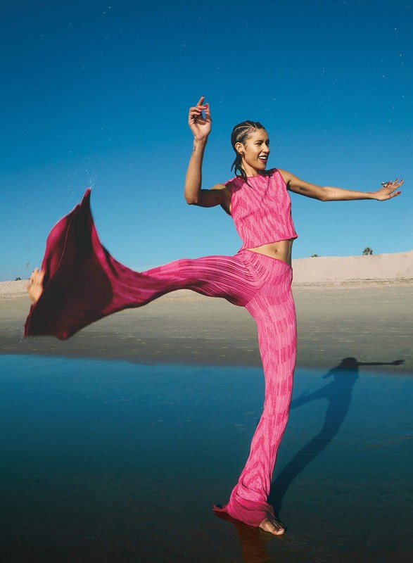 A young woman smiles and dances on a beach at dusk wearing a pink pleated two-piece outfit, with the sky and water in the background.