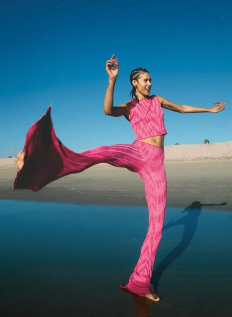 A young woman smiles and dances on a beach at dusk wearing a pink pleated two-piece outfit, with the sky and water in the background.