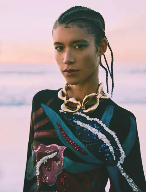 Portrait of a mixed race woman with braided hair wearing a statement gold necklace and patterned top, with a soft-focus coastal background.
