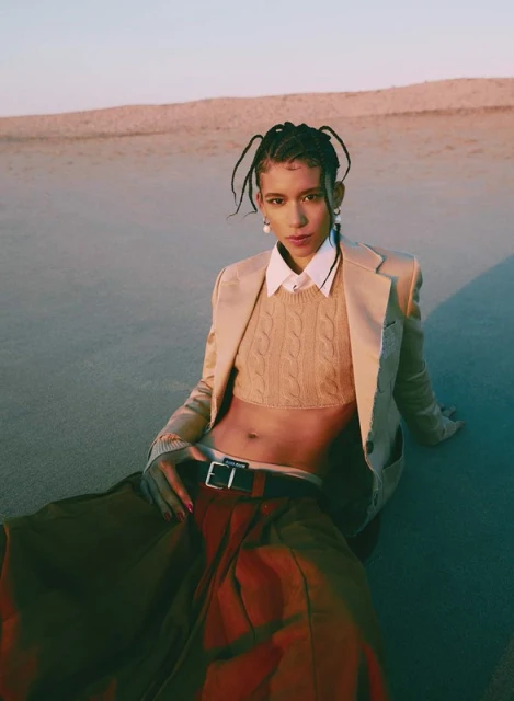 Woman in beige blazer and brown skirt sits on sand dune against sky in a fashion shot displaying her midriff, styled hair, and pearl earrings.