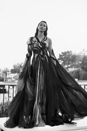 Model in a sweeping black gown with swirling sheer fabric panels standing outdoors against a bright sky background.