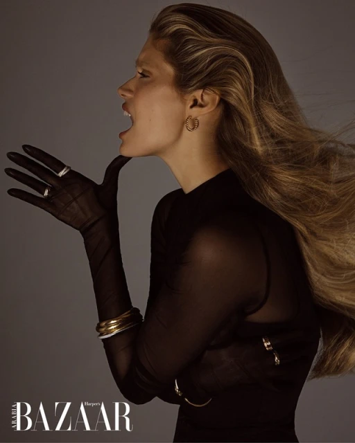 Profile shot of a blonde woman in a black top and gloves, posed with her mouth open against a grey background in a studio setting.