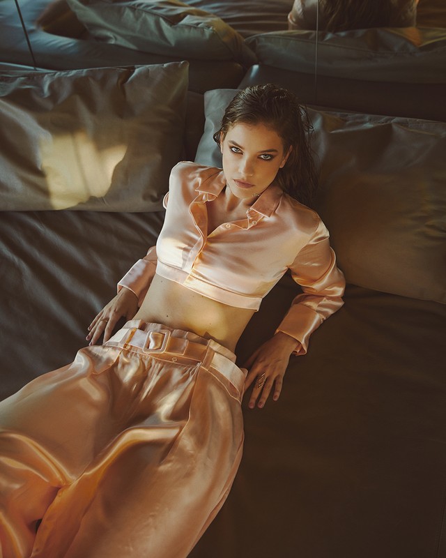 Brunette woman with wet hair laying on a bed with pillows in a shiny peach pajama set, looking at the camera in a studio setting.