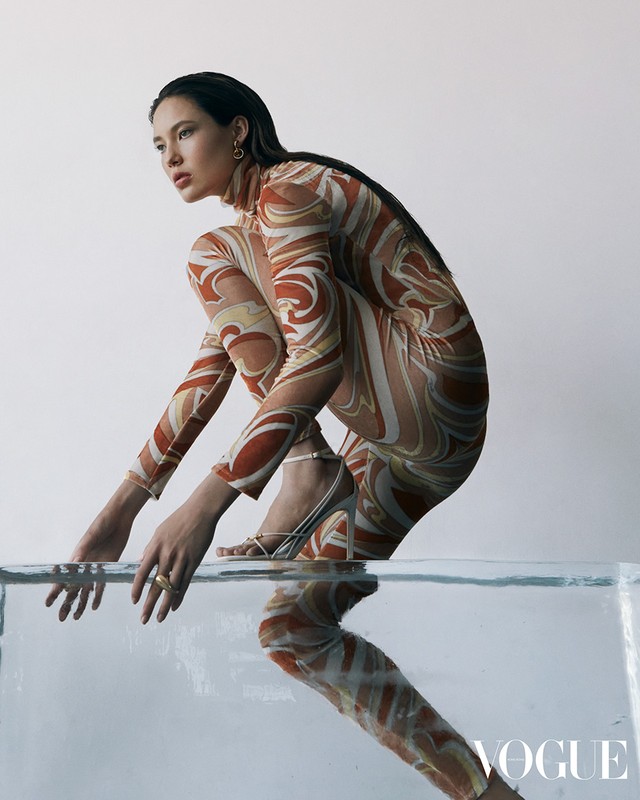 An adult woman in a patterned bodysuit poses on a translucent block in a studio, showcasing fashion and studio photography in a clean setting.