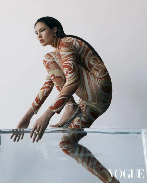 An adult woman in a patterned bodysuit poses on a translucent block in a studio, showcasing fashion and studio photography in a clean setting.