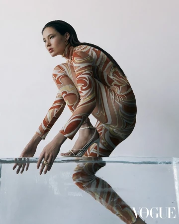 An adult woman in a patterned bodysuit poses on a translucent block in a studio, showcasing fashion and studio photography in a clean setting.