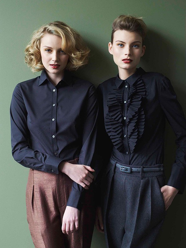 Two fair-skinned women pose indoors against a green wall. They both wear long-sleeve shirts and high-waisted pants in a fashion editorial.